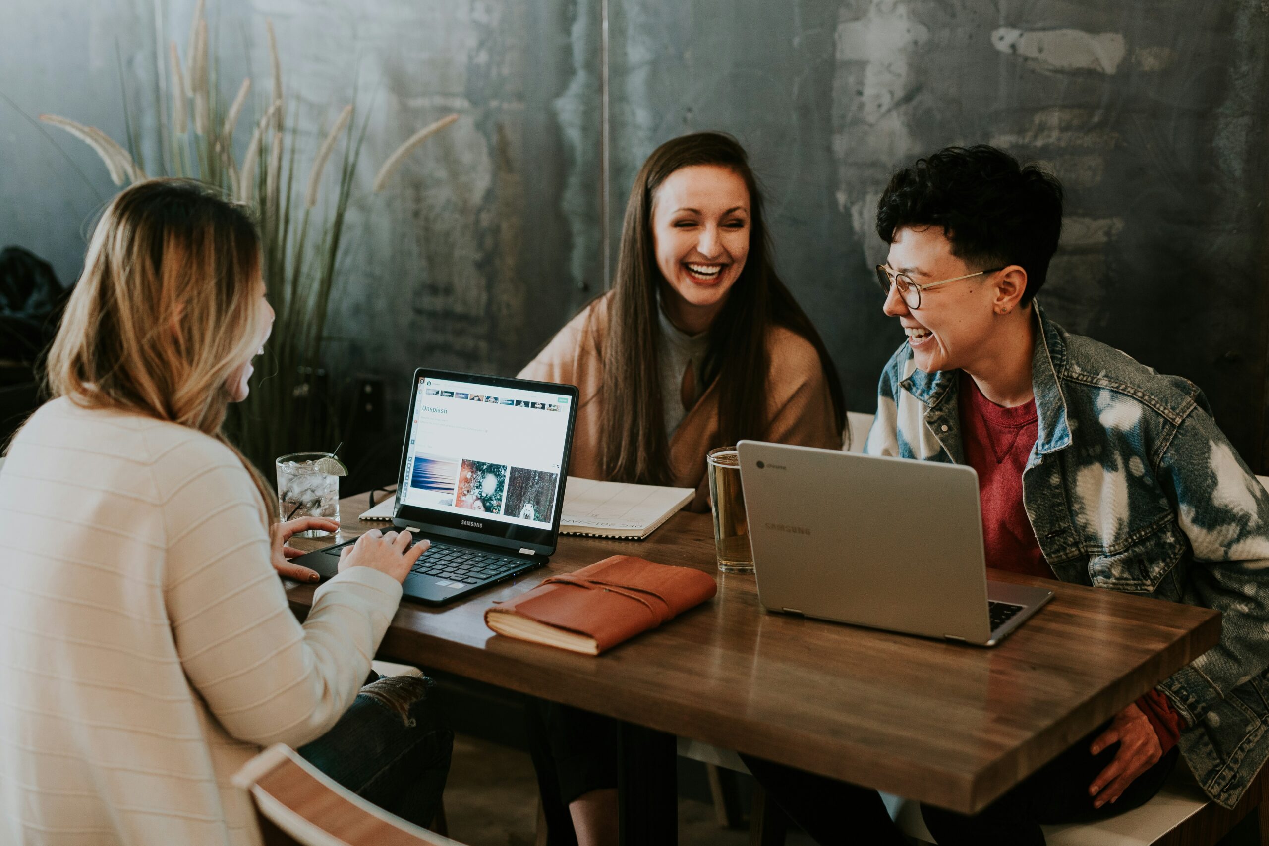 Três colegas de trabalho rindo e colaborando em um projeto com laptops em uma mesa de café.